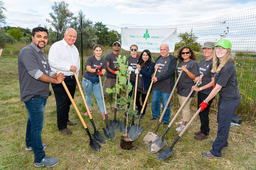 Group of people wearing National Tree Day t-shirts, holding shovels, standing around a young potted tree.