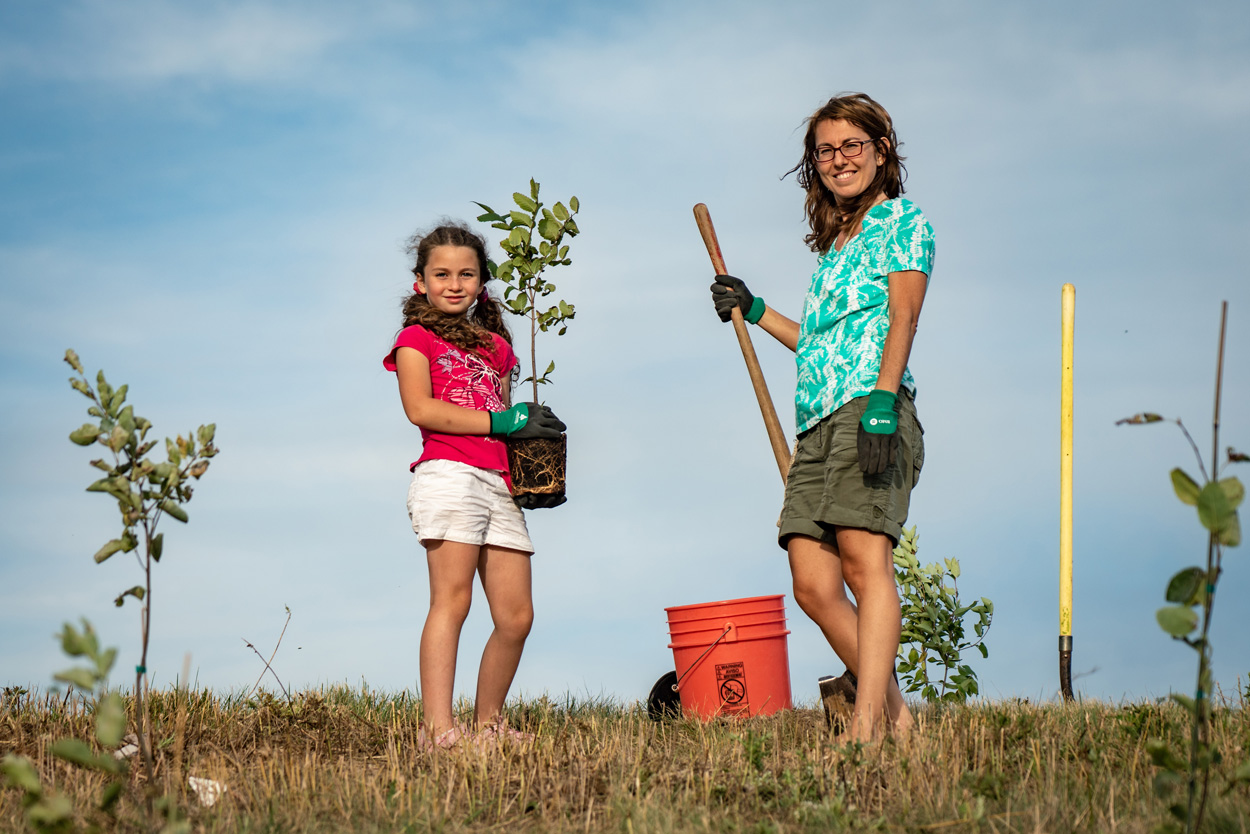 Young girl with mom posing for the camera, one holding a shovel and the other a young tree.