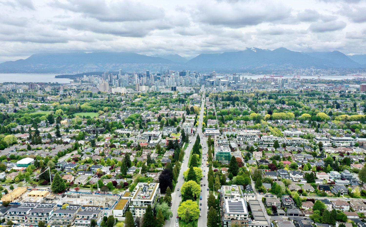 Photo de la ville de Vancouver vue d’en haut montrant des rues bordées d’arbres, des parcs, l’océan et les montagnes avec un ciel nuageux au-dessus.