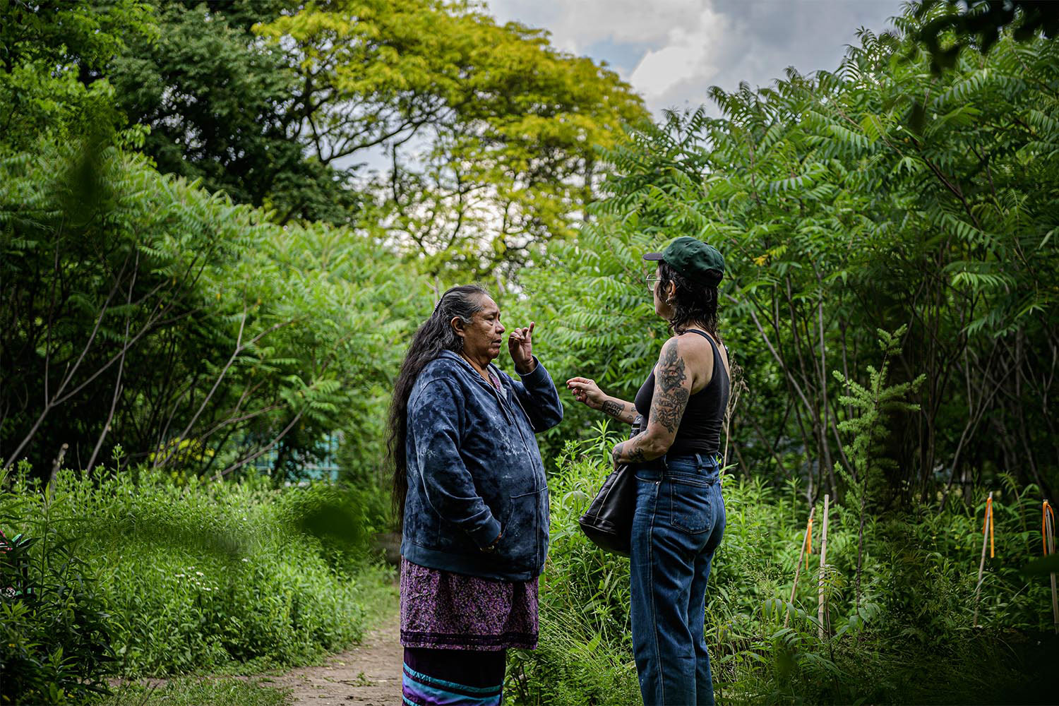 Une femme autochtone et une femme tatouée avec des lunettes se tiennent ensemble dans une forêt. On voit un ciel nuageux en arrière-plan.